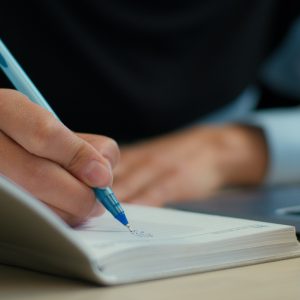 Photograph showing a person writing with a blue pen in a notebook placed on a desk next to a laptop.