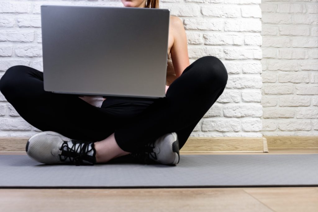 A woman in sportswear sitting on a yoga mat on the floor and using a laptop