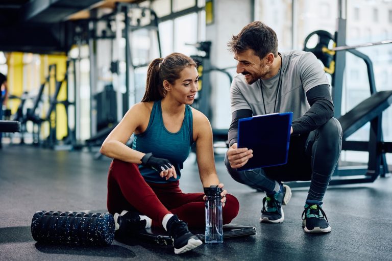 A man crouching down, holding a tablet, to talk to a woman sitting on the ground inside a gym