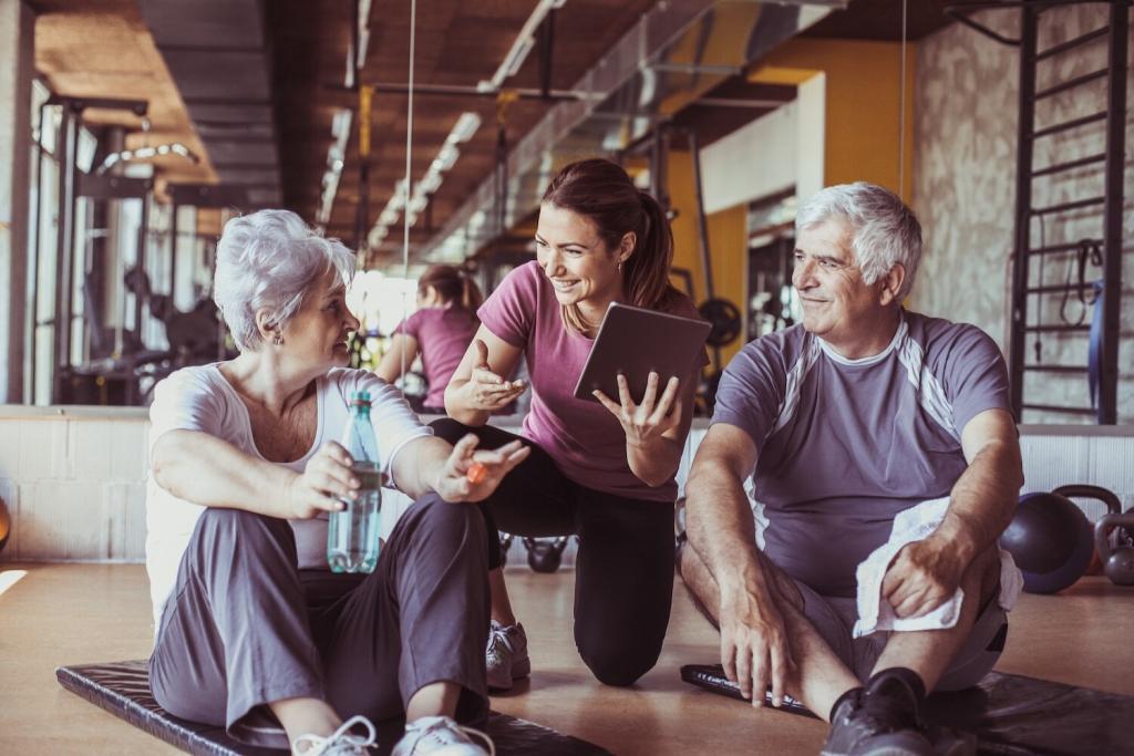 Senior couple in gym, with a personal trainer showing them something on digital tablet
