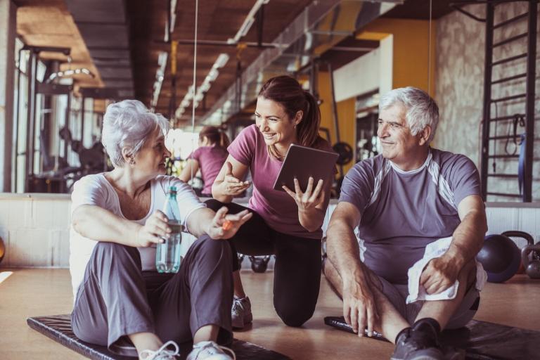 Senior couple in gym, with a personal trainer showing them something on digital tablet